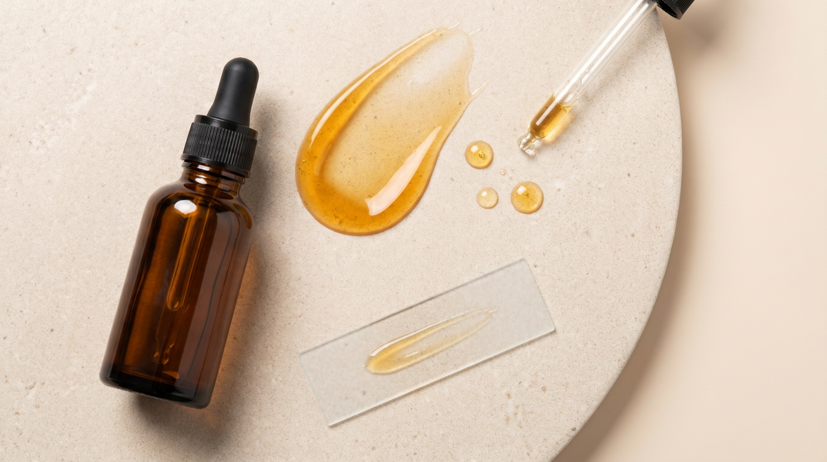 Flat-lay of an amber retinal serum dropper with a golden serum swatch, droplets, a glass pipette and a frosted slide on a stone surface
